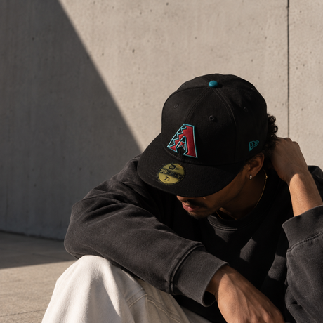 A person sitting outdoors against a concrete wall, wearing a black baseball cap with a red and teal Arizona Diamondbacks logo and a gold size 7 sticker on the brim, along with a dark sweatshirt and off-white pants.