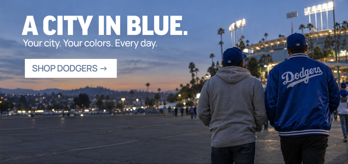 Two people seen from behind standing in a stadium at dusk, one wearing a gray hoodie and the other a blue Los Angeles Dodgers varsity jacket. Large text on the left reads 'A CITY IN BLUE.